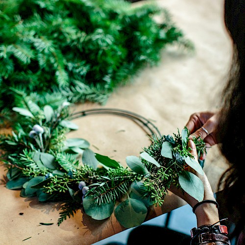 A handmade wreath being crafted with greenery and eucalyptus, by a person’s hands on a wooden table, with tools and foliage nearby.