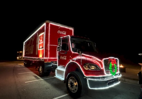 A bright red Coca‑Cola delivery truck at night, decorated with lights and a green wreath on the front grille, parked in a lot.