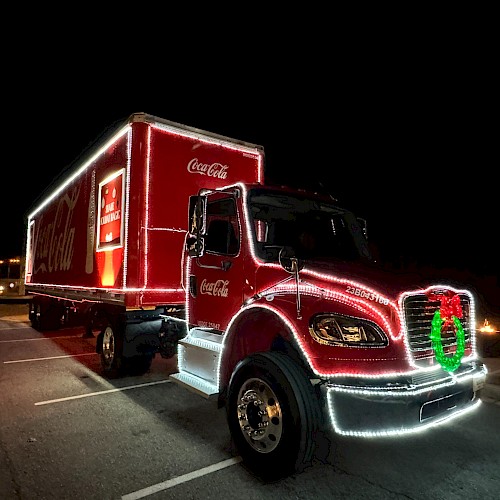 A bright red Coca‑Cola delivery truck at night, decorated with lights and a green wreath on the front grille, parked in a lot.