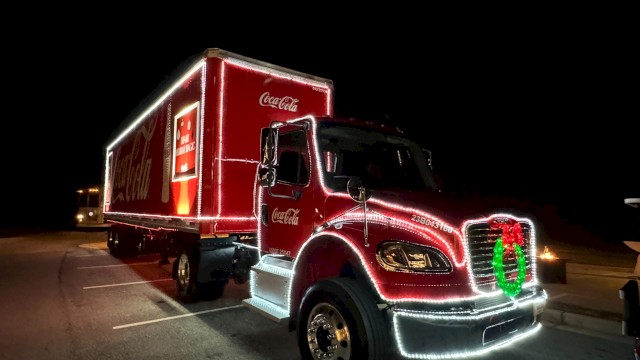 A bright red Coca‑Cola delivery truck at night, decorated with lights and a green wreath on the front grille, parked in a lot.