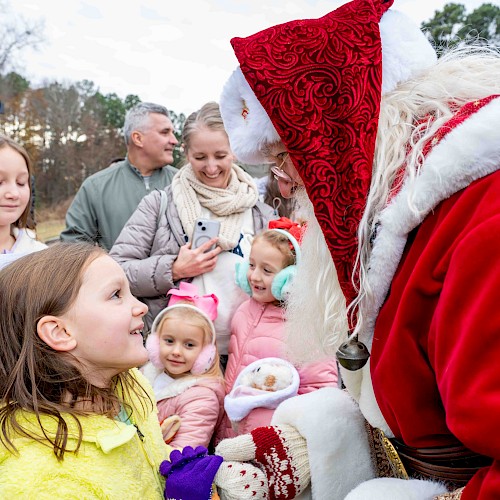 Santa Claus greets children outdoors, sharing a joyful moment during a festive event on a cloudy day.