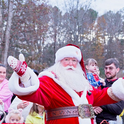 A cheerful Santa waves to a crowd in a park, with kids and adults gathered, festive outfits and a cozy holiday vibe. Ending this sentence.