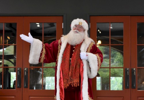 A person dressed as Santa Claus stands in front of wooden doors, waving with one hand in a red suit and white fur trim.