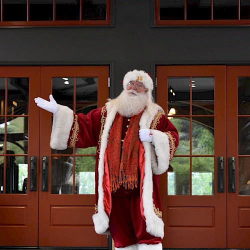A person dressed as Santa Claus stands in front of wooden doors, waving with one hand in a red suit and white fur trim.