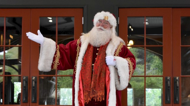 A person dressed as Santa Claus stands in front of wooden doors, waving with one hand in a red suit and white fur trim.