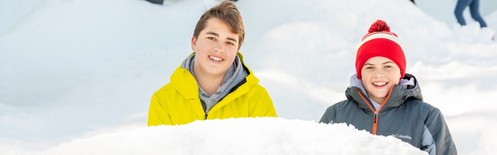 Two smiling kids in bright jackets sit in a snowy field with others playing and digging snow in the background.