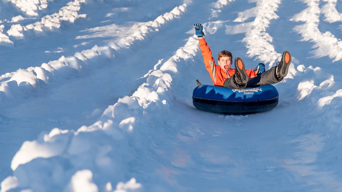 A child wearing an orange jacket rides a blue tube down a snowy slope, arms up in excitement, surrounded by fluffy white snow.