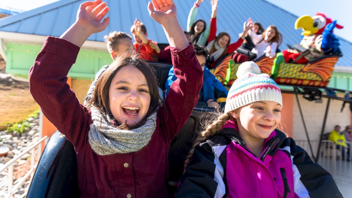 Two smiling kids on a roller coaster with raised arms, fast-tracked by cheering riders in the background aboard a colorful amusement ride.