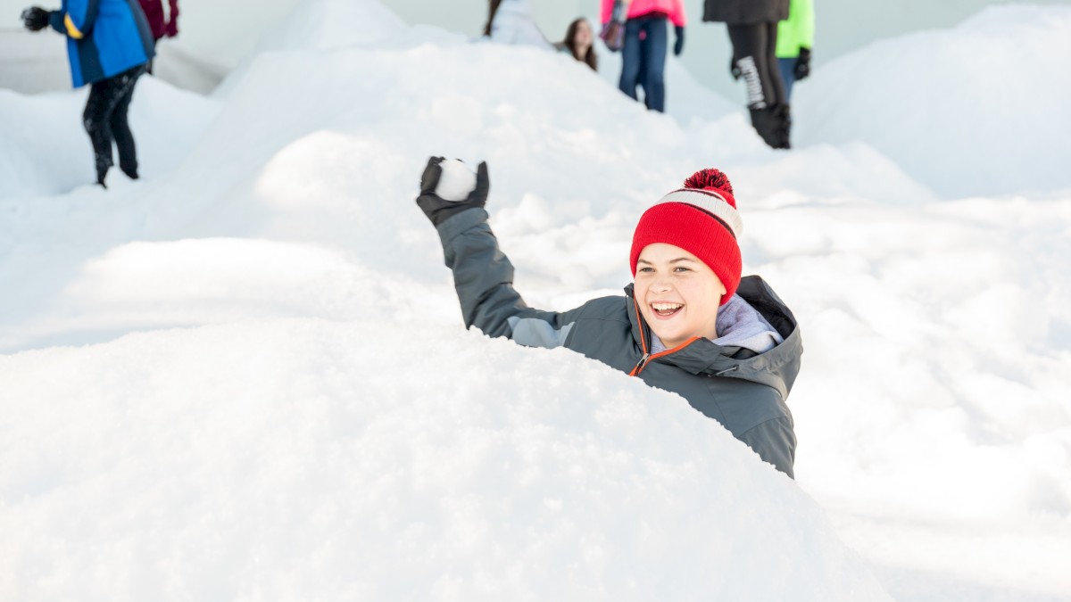Kids playing in a snowy landscape, one smiling child in a red hat waves from a snow pit while others stand and walk in the background.