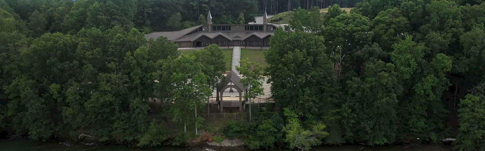 The image shows a large building complex surrounded by dense trees, situated near a body of water, possibly a lake or river.