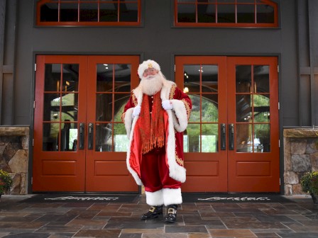 A Santa figure stands on a tiled porch in front of orange doors, wearing a red suit with white trim, black boots, and a red scarf, festive pumpkins nearby.