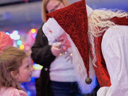 A child greets Santa Claus, who leans in for a warm, festive conversation at a colorful holiday event.