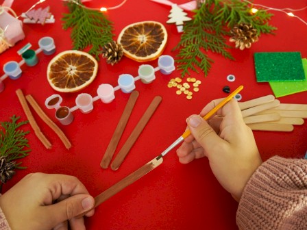 A cozy Christmas crafting scene: hands shaping sticks, dried orange slices, pine branches, red table, beads, and mini glittery decorations.