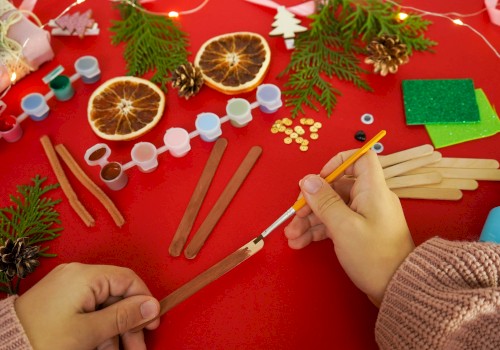 A crafting scene with cinnamon sticks, dried orange slices, pine sprigs, beads, and wooden sticks on a red table, hands shaping something festive.