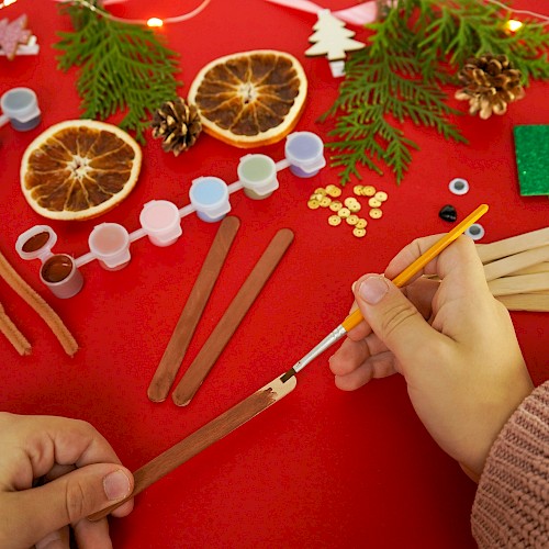 A crafting scene with cinnamon sticks, dried orange slices, pine sprigs, beads, and wooden sticks on a red table, hands shaping something festive.