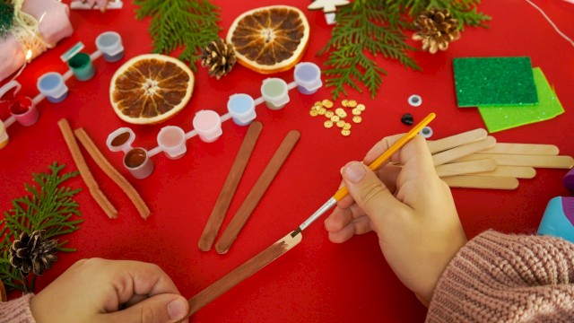 A crafting scene with cinnamon sticks, dried orange slices, pine sprigs, beads, and wooden sticks on a red table, hands shaping something festive.