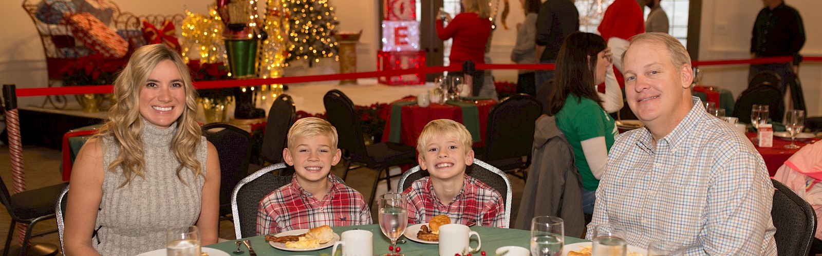 A family sits at a festive Christmas banquet table, smiling with decorated centerpieces and holiday treats, indoors with other guests in the background.