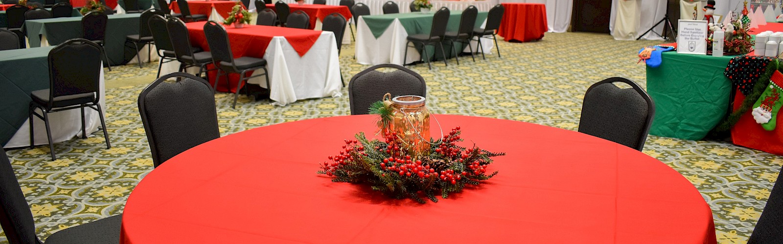 A banquet hall set for Christmas with round red tablecloths, chairs, a decorated central centerpiece, and a long head table in the background.