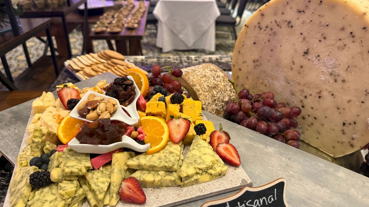 A cheese board display with assorted cheeses, fruits, nuts, crackers, and a wheel of cheese in the background, labeled “Artisan cheese display.”