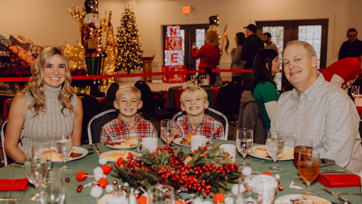 A family sits at a holiday table with festive decorations, poinsettias, and Christmas trees in the background, smiling for the camera.