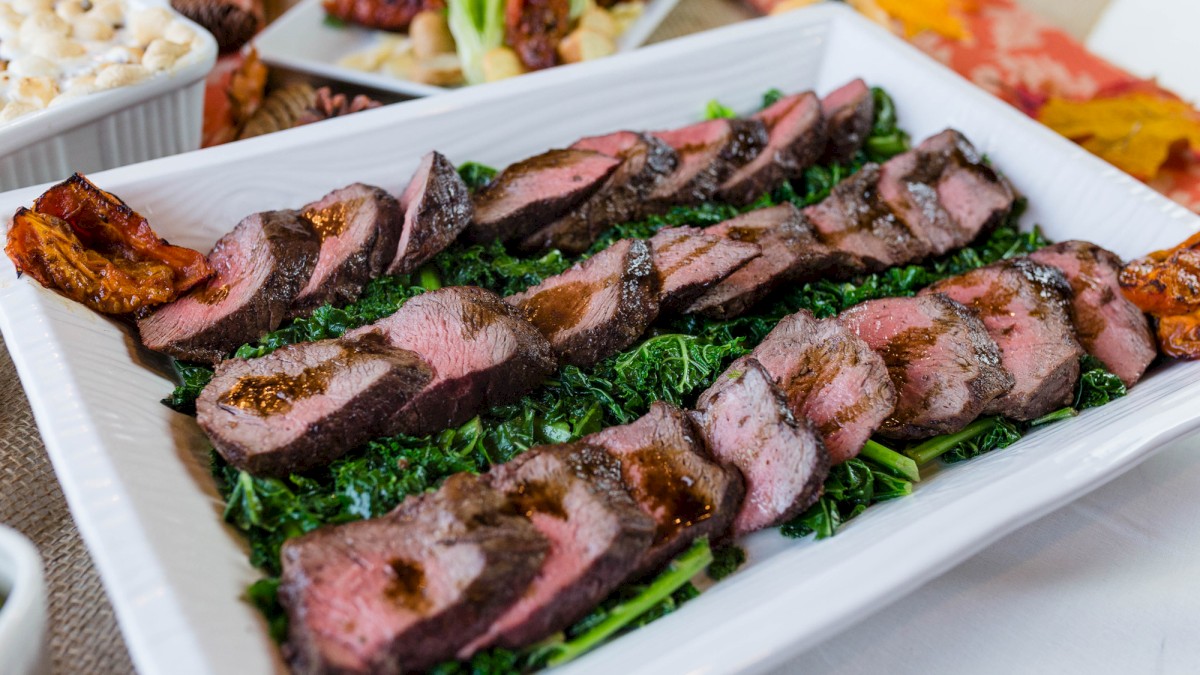 Grilled sliced steak on a bed of greens, juicy and medium-rare, served on a white platter with sides in the background.