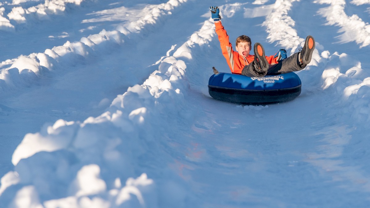 A child rides a blue snow tube down a snowy slope, arms raised in joy, surrounded by white, fluffy snow.