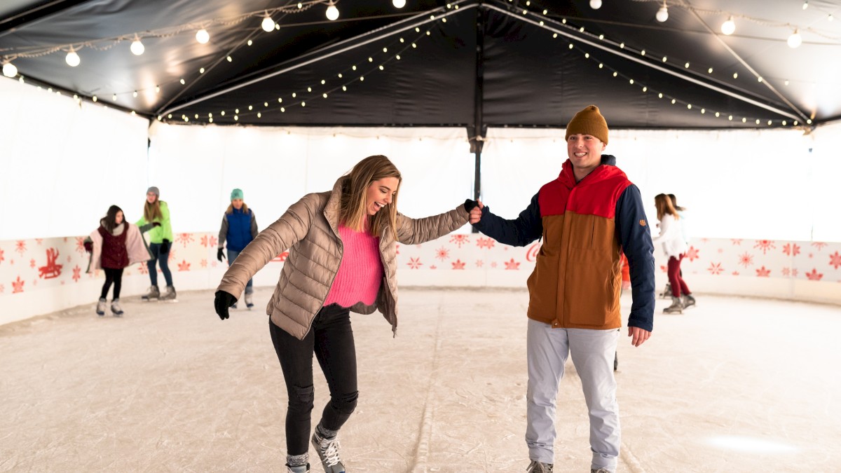 Two people skate hand-in-hand on an indoor rink while others figure skate in the background under string lights, enjoying a chilly day out.