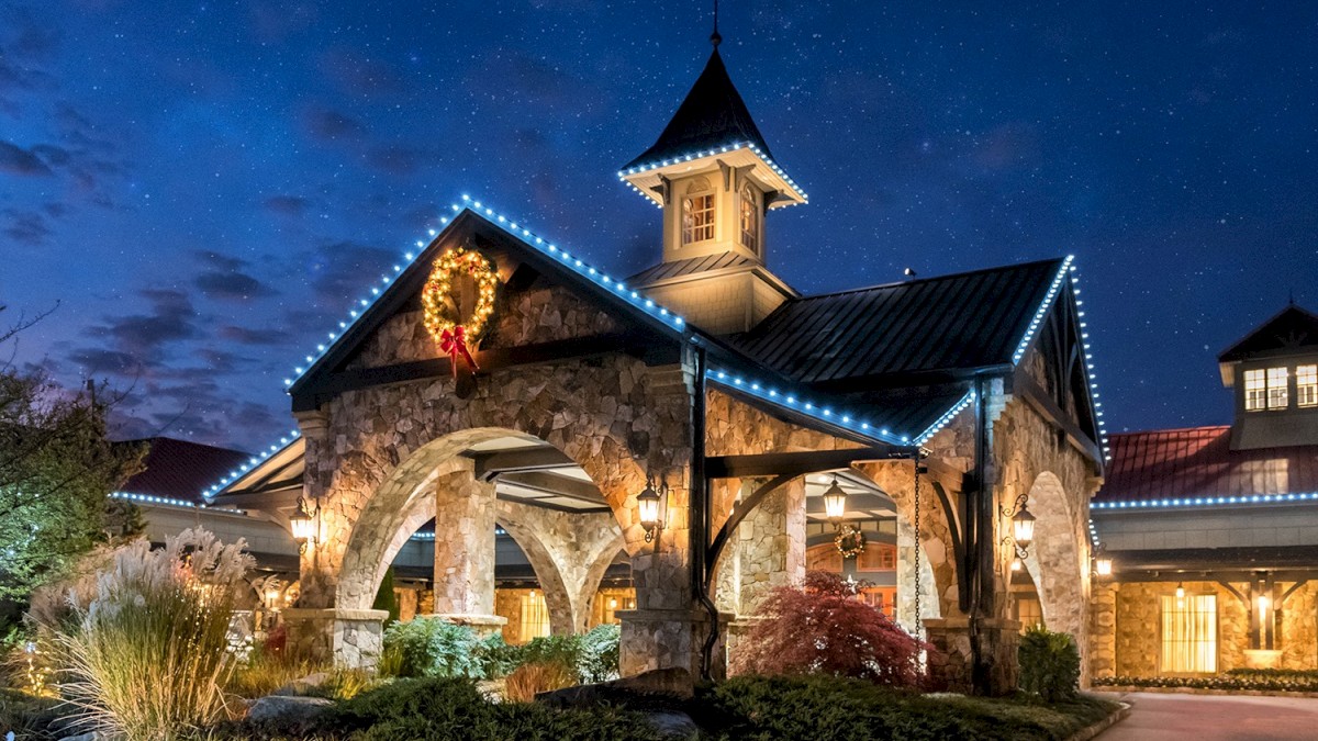 A charming stone building with a tall turret, festive wreath, and blue string lights outlining arches; dusk sky, inviting entryway.