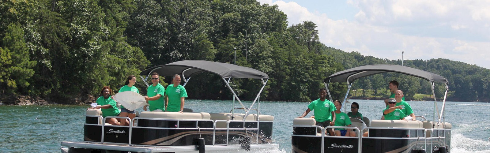Two pontoons with people wearing green shirts are speeding on a lake surrounded by trees under a partly cloudy sky.