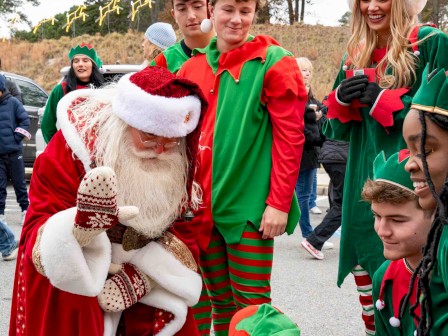Santa greets kids and elves in a festive outdoor parade; everyone smiles, wearing colorful Christmas outfits and hats.