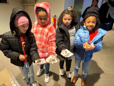 Four kids in hooded jackets are proudly holding small paper crafts or snacks they made, inside a room.