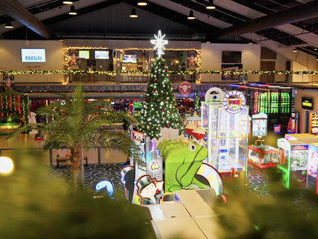 A festive arcade scene with a tall Christmas tree, colorful arcade machines, holiday lights, and games inside a shopping center.