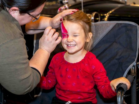 A makeup artist applies bright pink paint to a smiling young girl seated in a wheelchair.