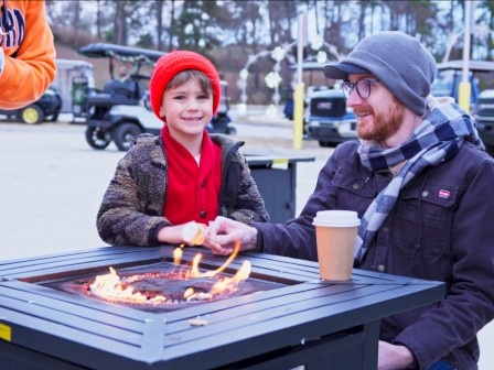 Two people sit by a fire pit table outdoors as a child and adult chat, coffee cup nearby, others stand around, winter clothing.