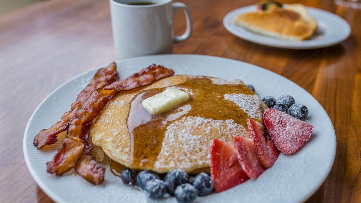 A plate with pancakes, butter, maple syrup, bacon, berries, and sliced fruit; a coffee mug and another pastry on the table in the background.