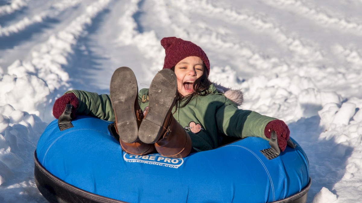 A joyful child in a green jacket and red hat rides a blue snow tube down a snowy slope, feet up and laughing, with snow all around.