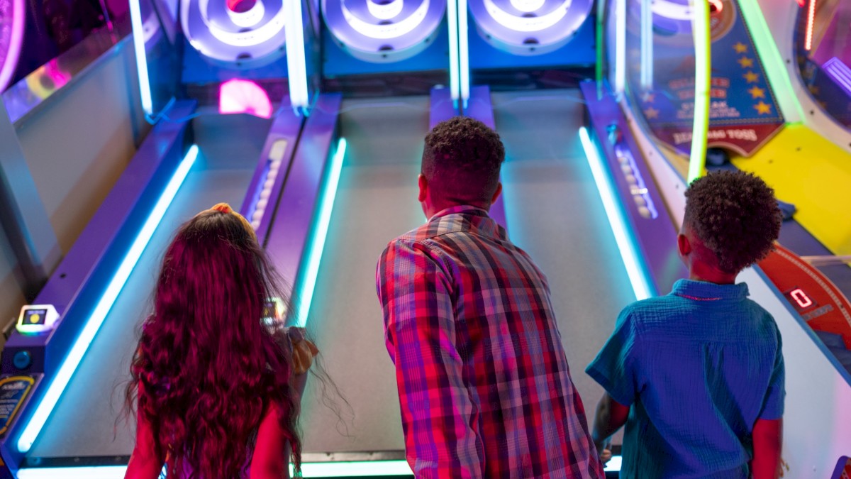 Three kids stand at a skee-ball/arcade lane, ready to roll balls under glowing blue lights in a colorful arcade.