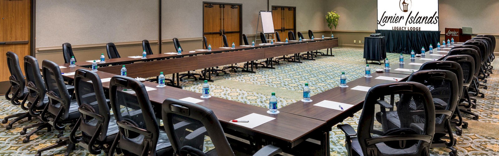 The image shows a conference room set up with a U-shaped table arrangement, chairs, water bottles, and writing materials. A screen displays 