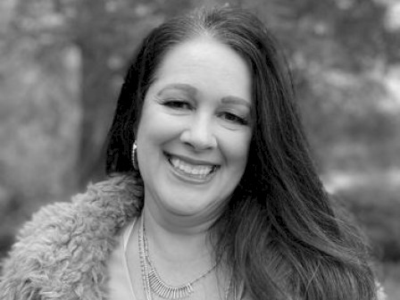 A smiling person with long hair is wearing a fur-lined coat and a necklace, standing outdoors with trees blurred in the background.