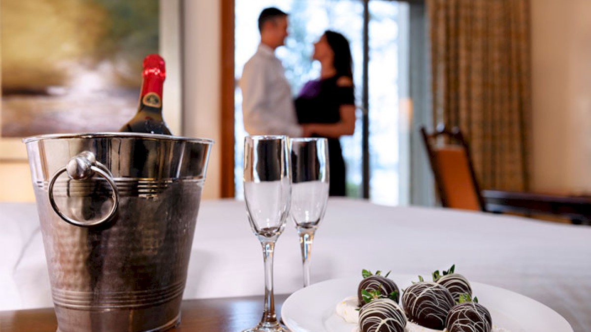 A champagne bottle chilling in a bucket, two fluted glasses, and a plate of elegantly decorated chocolate truffles on a table, with a blurred couple in the background.