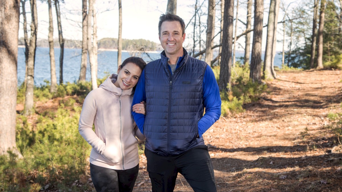 A couple walks arm-in-arm along a forest trail beside a lake, smiling, dressed in casual jackets, with trees and water in the background.