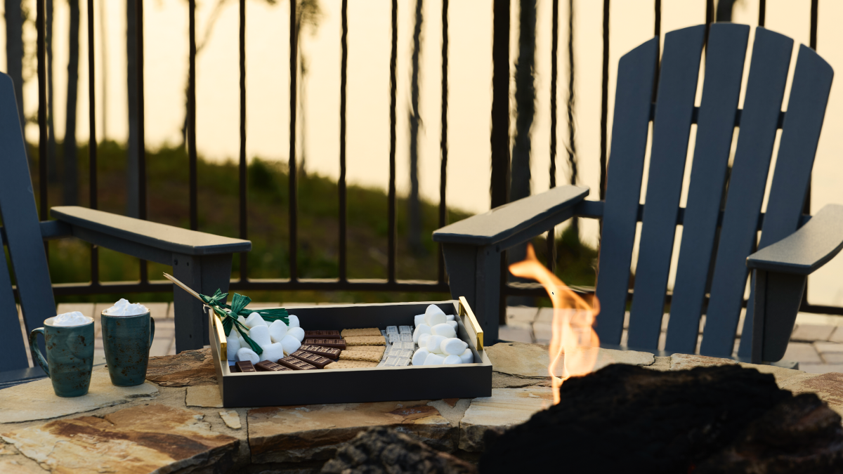 A cozy outdoor fire pit setup with popping flames, two blue chairs, marshmallows and goodies in a box, and drinks on a stone patio.
