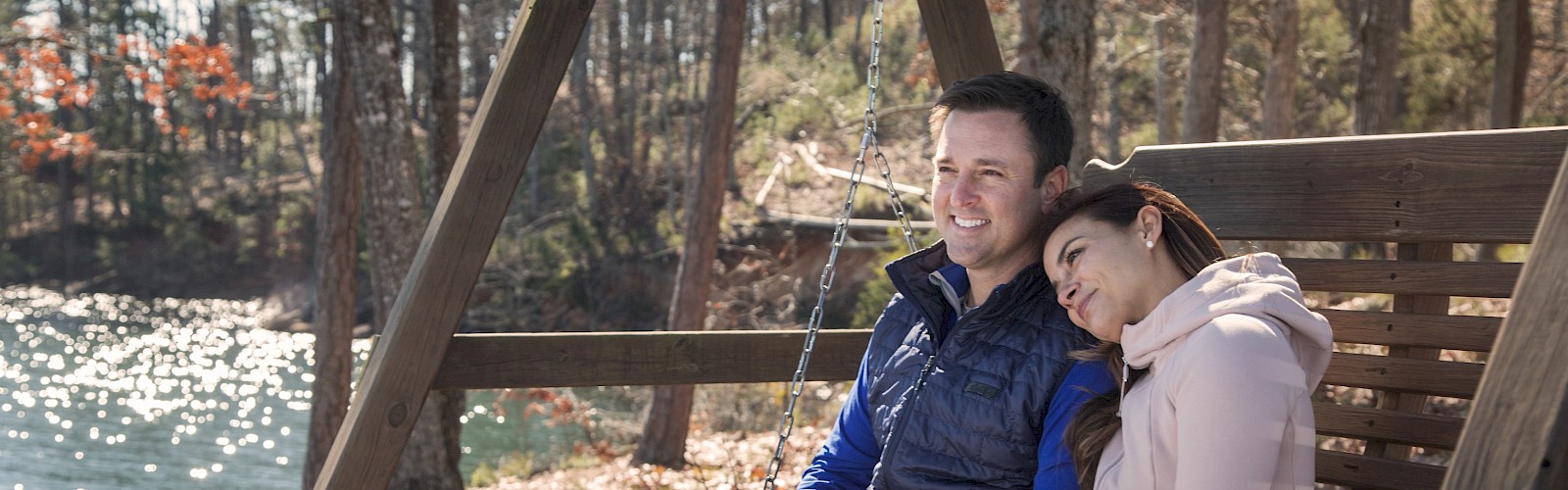 Two people sit close on a wooden swing by a sunny lake in a forest, smiling and relaxing together.