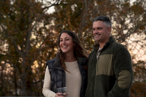 A smiling couple outdoors at dusk, the woman holding a cup while the man stands beside her, trees softly lit in the background.