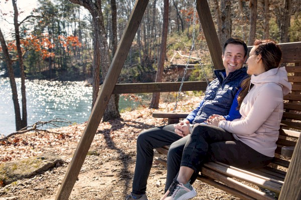 A couple sits on a wooden swing by a sunny lakeside, enjoying a chat and the autumn scenery.