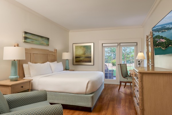 A clean hotel room with a king bed, two nightstands with lamps, a dresser, a chair, framed art, and French doors to a balcony, all on wooden floors.