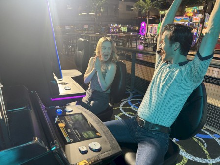 Two people celebrate at an arcade or driving simulator, one raising arms in excitement while the other looks surprised.