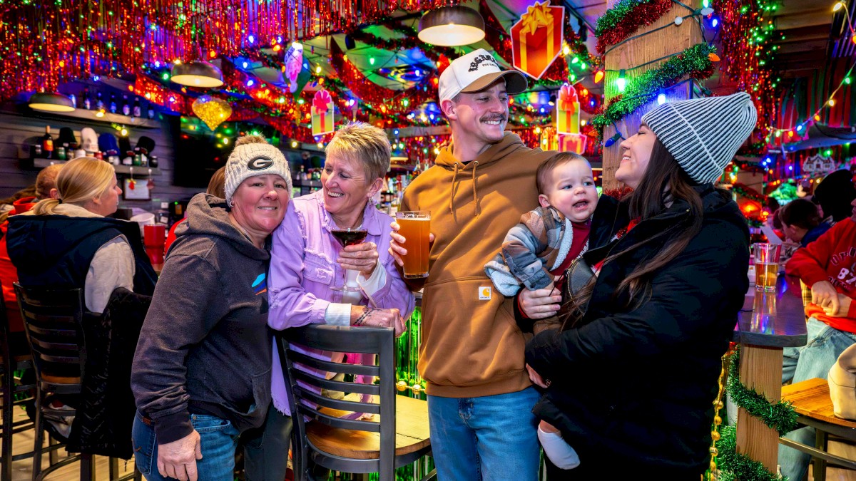 A festive bar scene with smiling adults and a child, colorful lights, garlands, and drinks, everyone enjoying a lively holiday party.