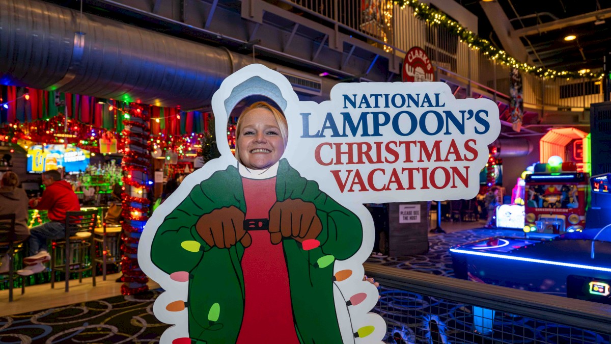 A festive indoor scene with colorful holiday lights, a cutout figure of a person in a green coat, and a sign reading “National Lampoon's Christmas Vacation.”