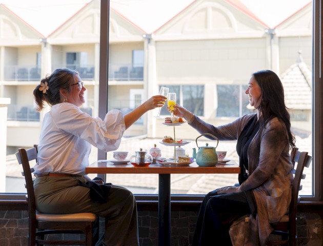 Two women sharing a friendly tea/pastry toast at a cafe by a large window, raising their cups in a cheerful moment.